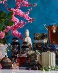 A colorful arrangement featuring a variety of organic tea products displayed on a table. The scene includes teapots, tea canisters, packets of Magic Hour's Goddess of Earl: Madagascar Vanilla Creme Tea for Soothing Delight & Delicious Decadence, a glass of tea, and a glass pitcher of iced tea with a star-shaped tea infuser. Pink cherry blossoms and a Buddha statue adorn the background.