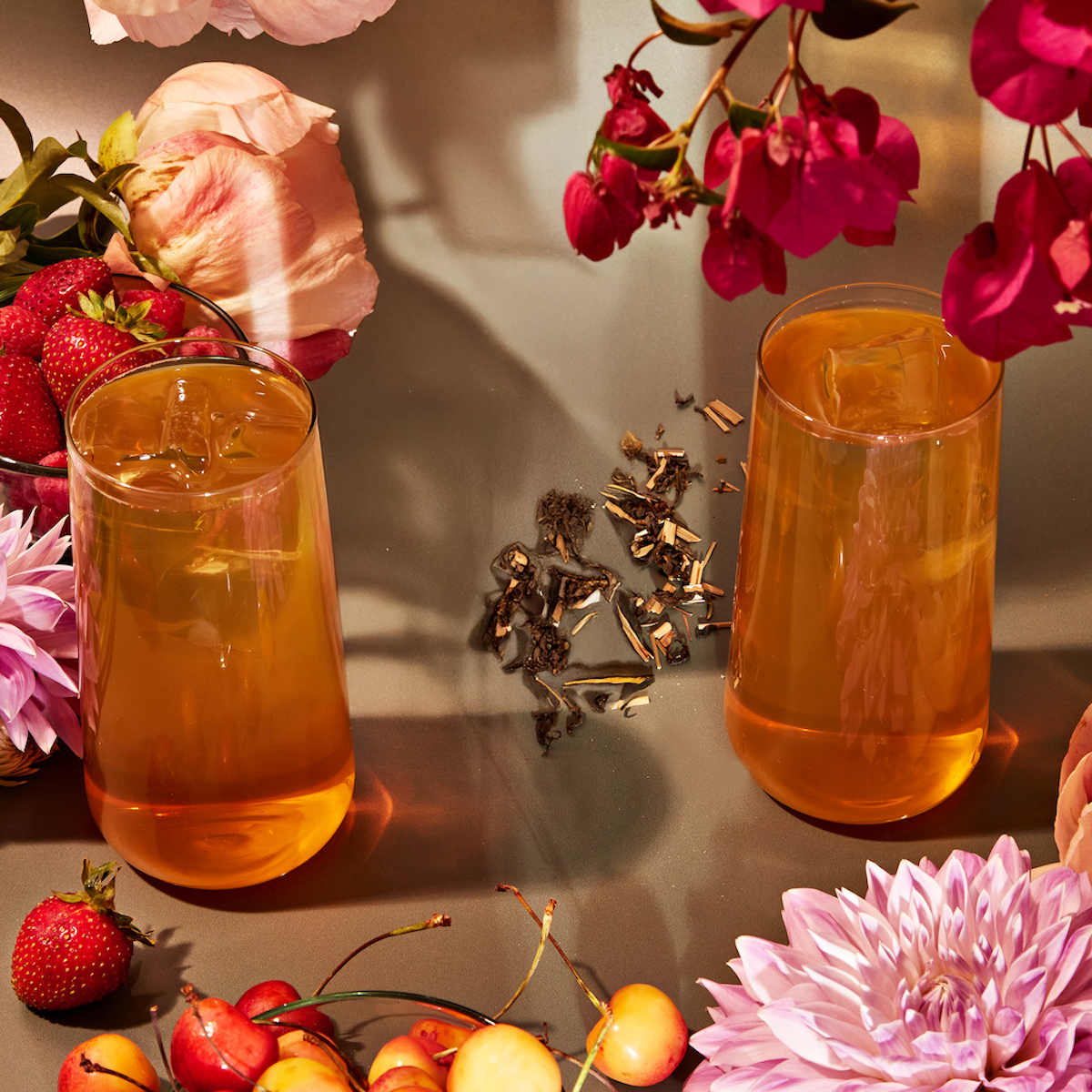 Two glasses of iced tea made with Goddess Green: Apricot Lychee Green Tea Bags are surrounded by fresh strawberries, cherries, pink peonies, dahlias, magenta flowers, and loose organic green tea leaves on a reflective surface.
