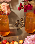 Two glasses of iced tea made with Goddess Green: Apricot Lychee Green Tea Bags are surrounded by fresh strawberries, cherries, pink peonies, dahlias, magenta flowers, and loose organic green tea leaves on a reflective surface.