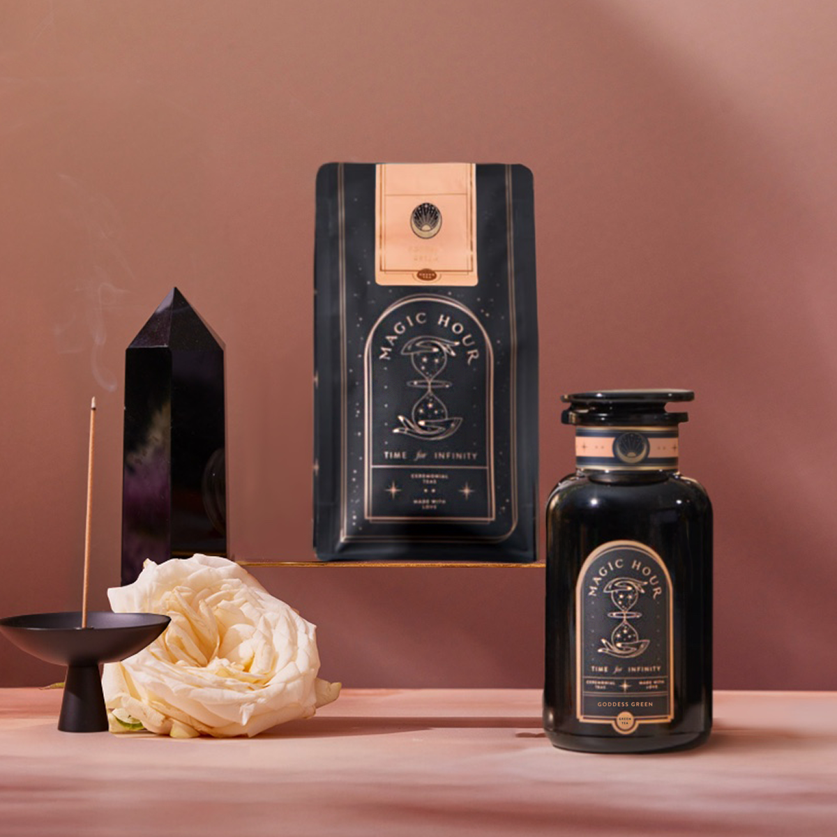 A Goddess Green Tea Starter Kit sits on a pink surface with a white rose, a burning incense stick in its holder, and a black crystal prism against a soft pink-brown backdrop.