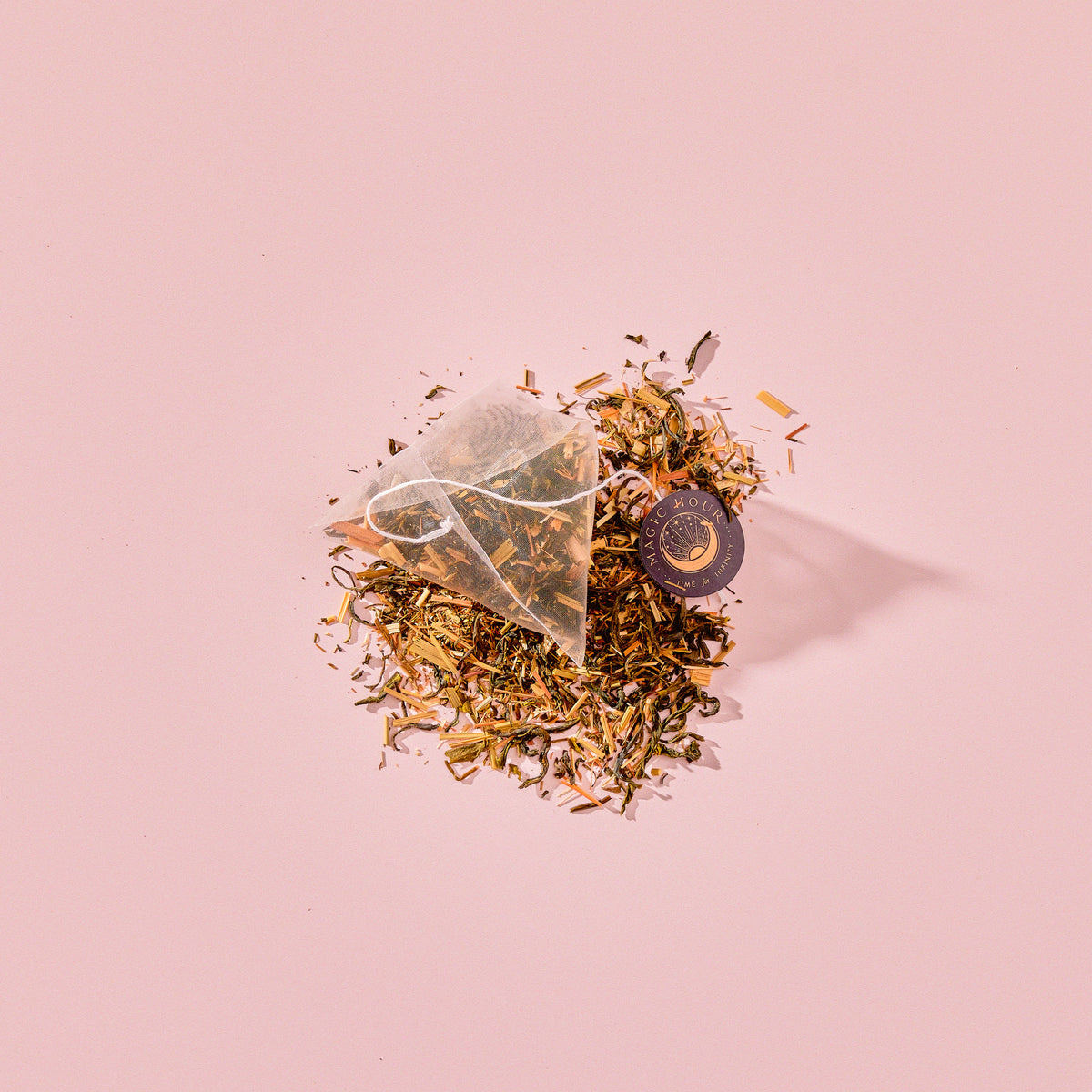 A pyramid-shaped green tea sachet of Tea for Abundance: Goddess Green: Apricot &amp; Lychee Green Tea with organic lemongrass resting on a pile of loose-leaf tea with a small circular tag.