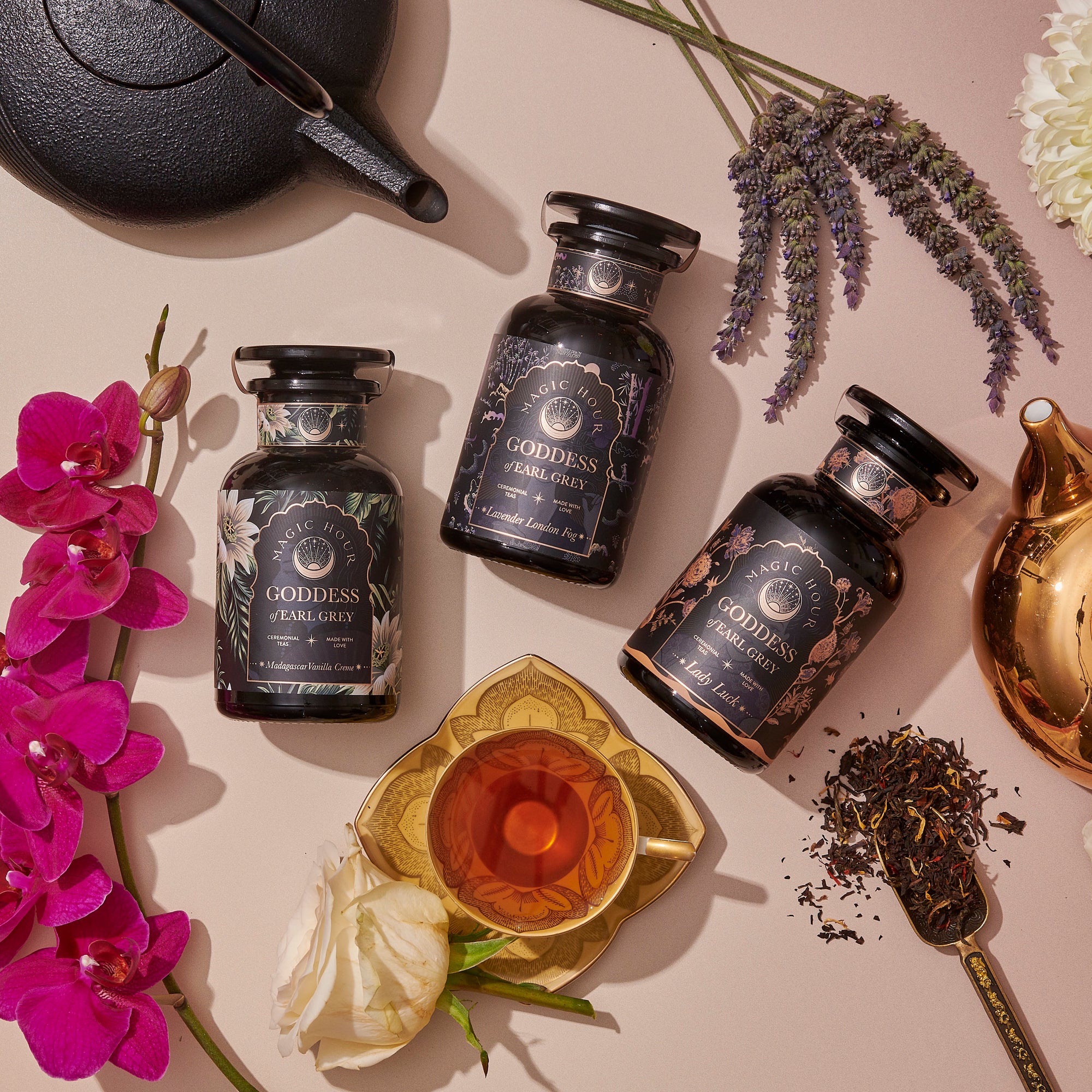 The Goddess of Earl: Lavender London Fog—Tea for Blooming Clarity &amp; Calm Moods sits in a labeled jar beside a glass teacup, loose tea leaves, a gold and black teapot, and flowers including an orchid and rose on a light surface.