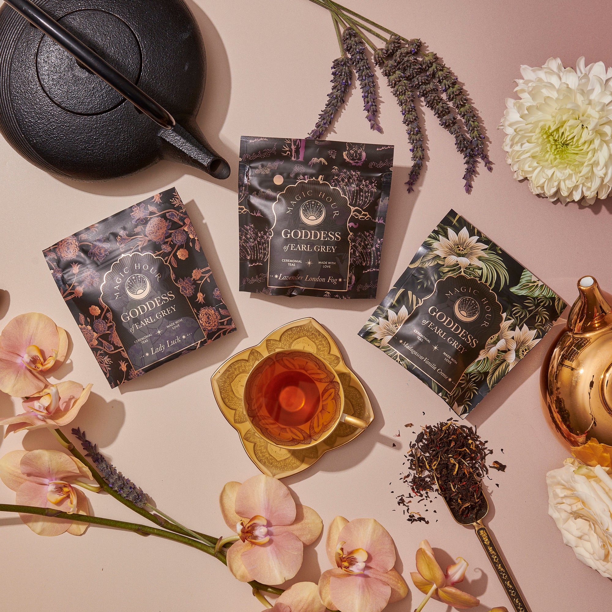 A flat lay of Goddess of Earl: Madagascar Vanilla Creme Tea packets, a teacup, black and gold teapots, loose organic black tea, and assorted flowers like orchids, lavender, and chrysanthemums on a soft pink surface.