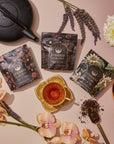 A flat lay of Goddess of Earl: Madagascar Vanilla Creme Tea packets, a teacup, black and gold teapots, loose organic black tea, and assorted flowers like orchids, lavender, and chrysanthemums on a soft pink surface.