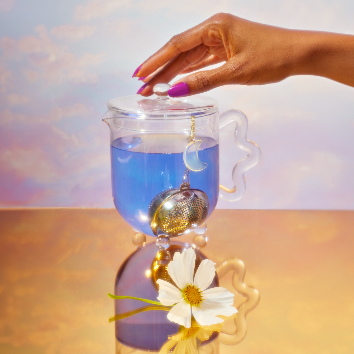 Blue tea in a glass teapot with a tea strainer hang off the side. A hand is grabbing the teapot lid. 