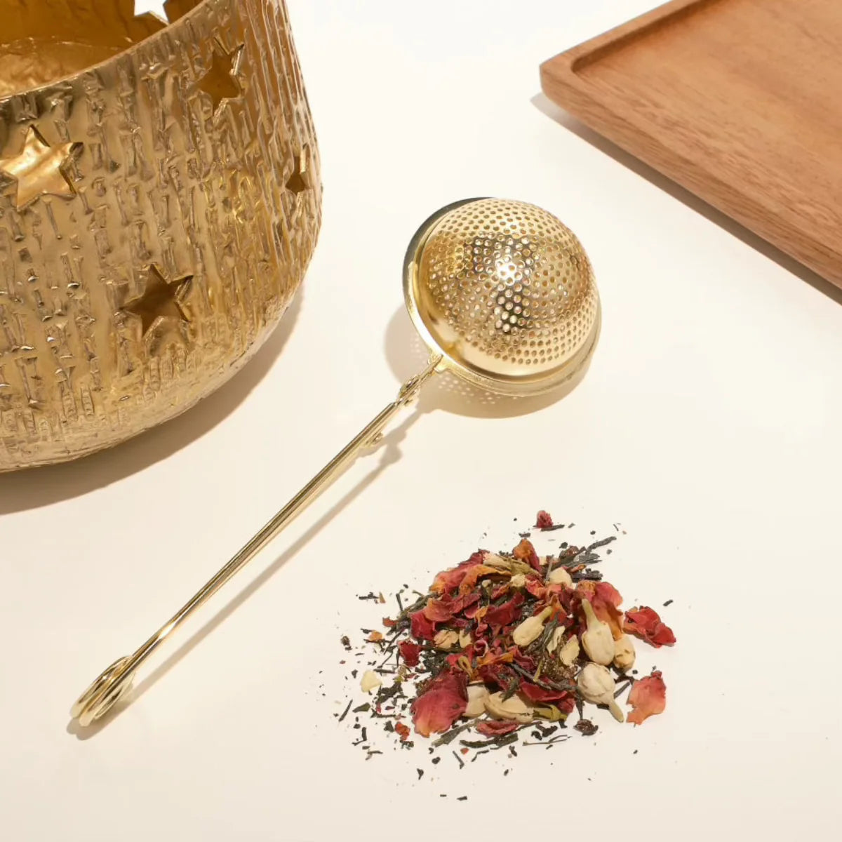 A gold tea infuser lies next to Tarot-Inspired Tea, adorned with rose petals and herbs. A decorative gold-colored jar with star cutouts is slightly visible on the left, all set against a wooden tray backdrop. Product shown: 12 Tarot Tea Traveler Jar Bundle.