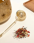 A golden tea infuser with a round mesh end sits on a white surface beside Earl Grey-inspired loose teas with rose petals. In the background, there's a textured gold pot with star cutouts and a wooden tray, capturing the essence of the London Fog Tea Sampler Set experience.