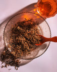 A clear glass plate holds a heap of Organic Golden Monkey Tip Yunnan Black Tea leaves from Magic Hour, accompanied by a wooden spoon resting in the leaves. To the top right, a translucent cup filled with amber-colored tea sits on the table, casting a warm glow. The background is a light pinkish hue.