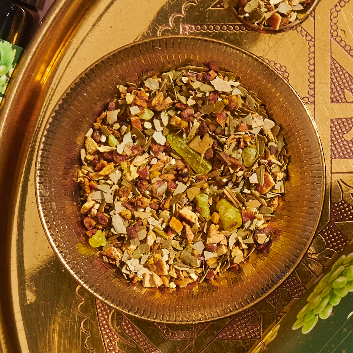 A close-up of a glass bowl filled with a colorful mixture of dried herbs, fruits, and spices. The bowl is set on an ornate, decorative golden tray with intricate patterns. The blend, reminiscent of Green Grape Yerba Mate: Cosmic Garden Iced Tea by Magic Hour, includes green, brown, and orange bits, indicating various ingredients.