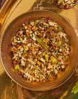 A close-up of a glass bowl filled with a colorful mixture of dried herbs, fruits, and spices. The bowl is set on an ornate, decorative golden tray with intricate patterns. The blend, reminiscent of Green Grape Yerba Mate: Cosmic Garden Iced Tea by Magic Hour, includes green, brown, and orange bits, indicating various ingredients.