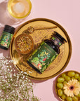 A golden tray holds a green bottle and a small jar with botanical labels, a bowl of loose leaf tea, and a golden spoon. Next to the tray are a bunch of grapes, a gerbera daisy, white flowers, and a glass with a round ice cube on a pink background. Perfect for your Magic Hour Green Grape Yerba Mate: Cosmic Garden Iced Tea experience.