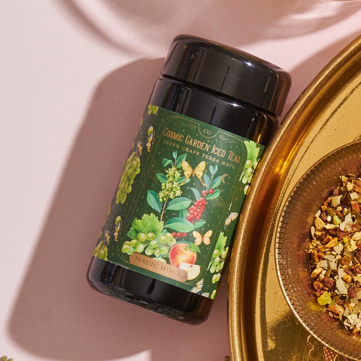 A black cylindrical container labeled &quot;Green Grape Yerba Mate: Cosmic Garden Iced Tea&quot; from Magic Hour is placed next to a gold dish filled with organic loose leaf tea leaves and herbs. The backdrop is a light pink surface with shadows adding depth.