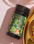 A black cylindrical container labeled "Green Grape Yerba Mate: Cosmic Garden Iced Tea" from Magic Hour is placed next to a gold dish filled with organic loose leaf tea leaves and herbs. The backdrop is a light pink surface with shadows adding depth.