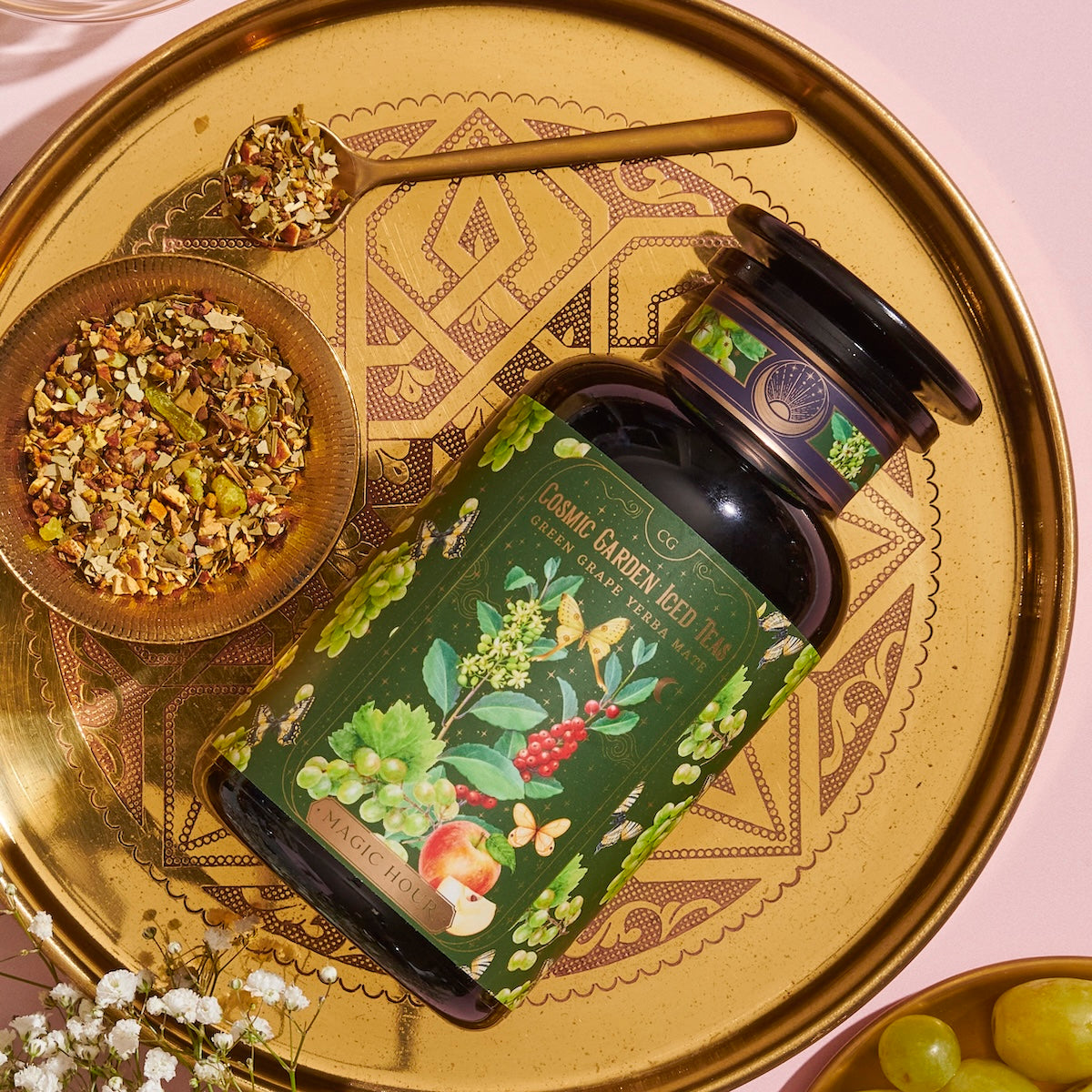 A decorative gold tray holds a dark green bottle of organic tea labeled &quot;Green Grape Yerba Mate: Cosmic Garden Iced Tea&quot; by Magic Hour, with images of flowers, herbs, and fruits. The tray also has a small wooden bowl filled with loose tea leaves and a gold spoon resting beside it.
