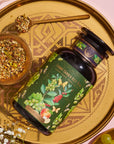 A decorative gold tray holds a dark green bottle of organic tea labeled "Green Grape Yerba Mate: Cosmic Garden Iced Tea" by Magic Hour, with images of flowers, herbs, and fruits. The tray also has a small wooden bowl filled with loose tea leaves and a gold spoon resting beside it.