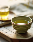 A ceramic cup of Guizhou green tea rests on a wooden tray, next to a small glass cup of Organic Decaf Green Tea and a folded napkin, all softly illuminated in the background.
