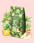 A package of Green Grape Yerba Mate: Cosmic Garden Iced Tea features grape, apple, pineapple, and leaf illustrations on a pink background; fresh grapes, sliced pineapple, apple, green leaves, and vibrant turmeric root are arranged around the package.