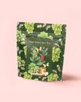 A resealable green pouch labeled "Green Grape Yerba Mate: Cosmic Garden Iced Tea" features grape, flower, bee, and fruit illustrations with a touch of spirulina on a pink background.