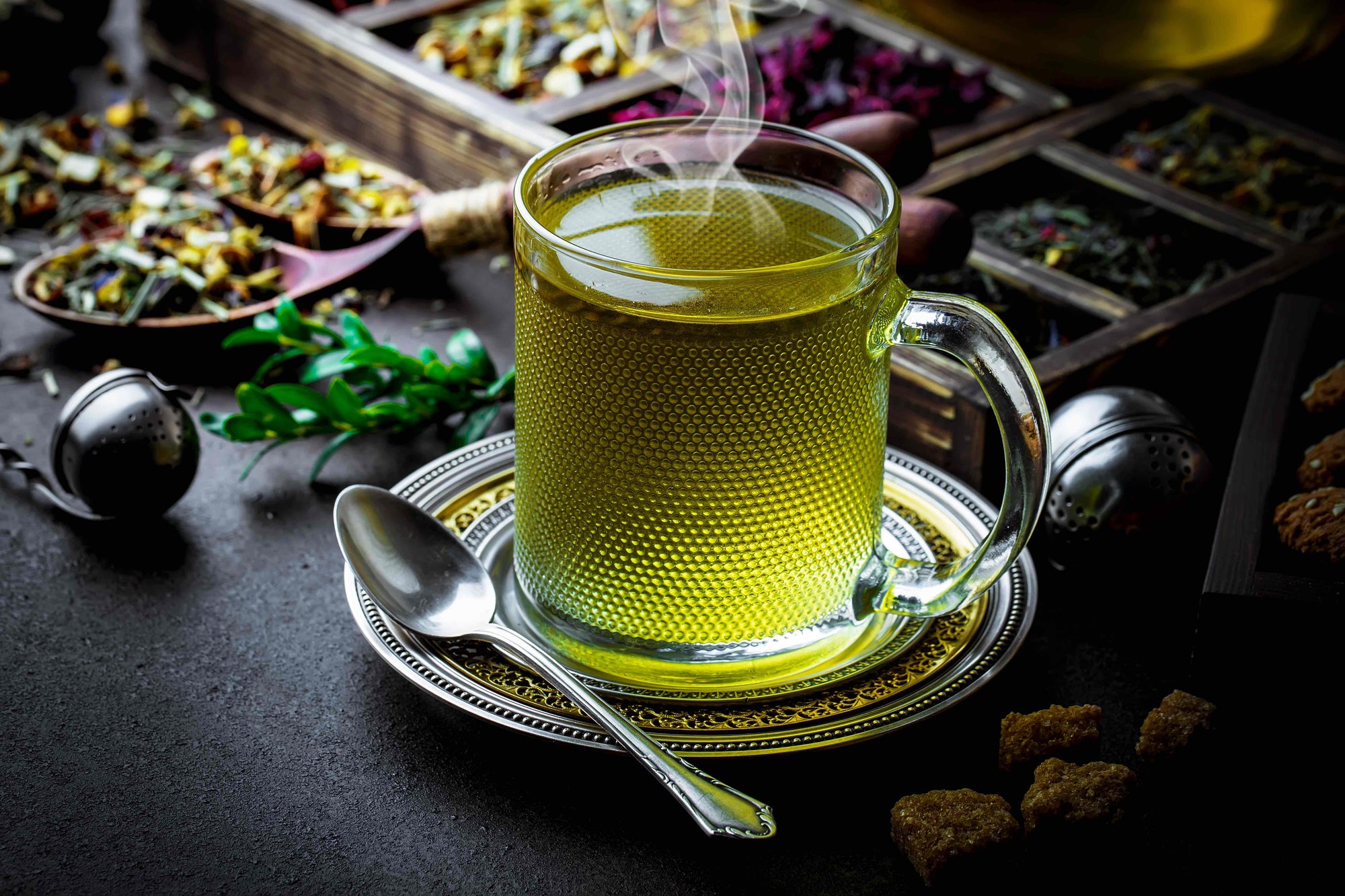 A clear glass cup of steaming green tea sits on a decorative saucer with a silver spoon by its side. Surrounding the cup are various compartments of loose tea herbs and spices, a tea infuser, and brown sugar cubes on a dark background.