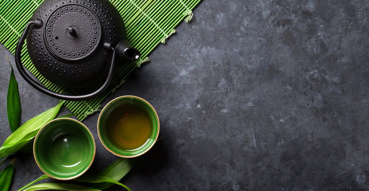 A black cast iron teapot is placed on a green bamboo mat, with two green teacups filled with tea beside it. The set is positioned on a dark slate surface, accompanied by green leaves, creating a serene and minimalist composition.