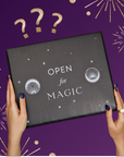 A person with dark blue nails holds the Black Friday Mystery Box, labeled "OPEN FOR MAGIC," against a purple background with gold stars, fireworks, and question marks—perfect for discovering premium teas and unique tea accessories.