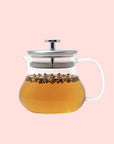The Essential Perfection Teapot with Built-in Strainer, made from borosilicate glass and featuring a metal lid, is filled with light brown tea and loose tea leaves and set against a pale pink background.