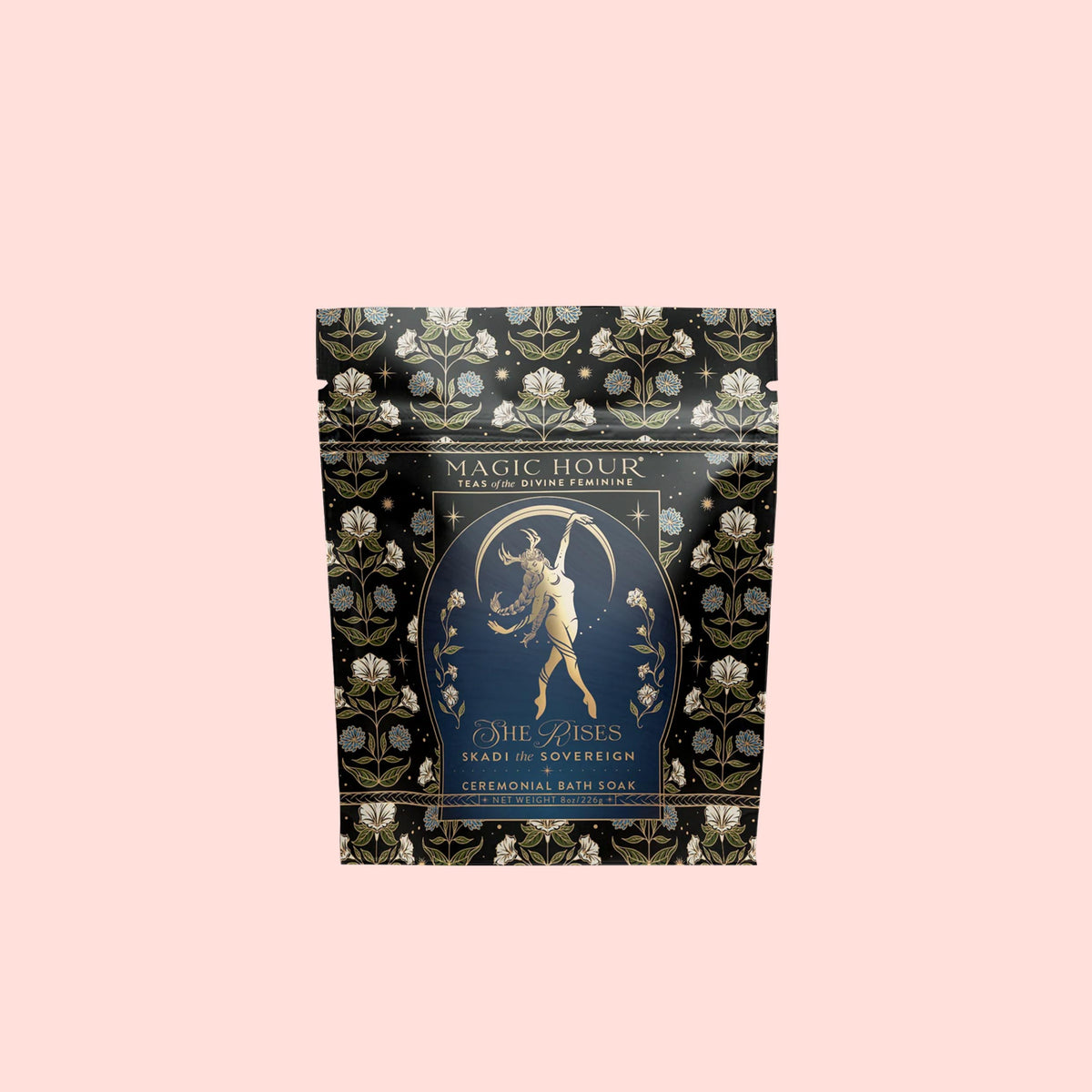 A black and gold floral Magic Hour bath tea sachet labeled “Skadi The Sovereign” features an illustration of a woman holding a staff in a celestial design. This botanical soak stands against a pale pink background, inviting you to indulge in its enchanting winter bath blend.