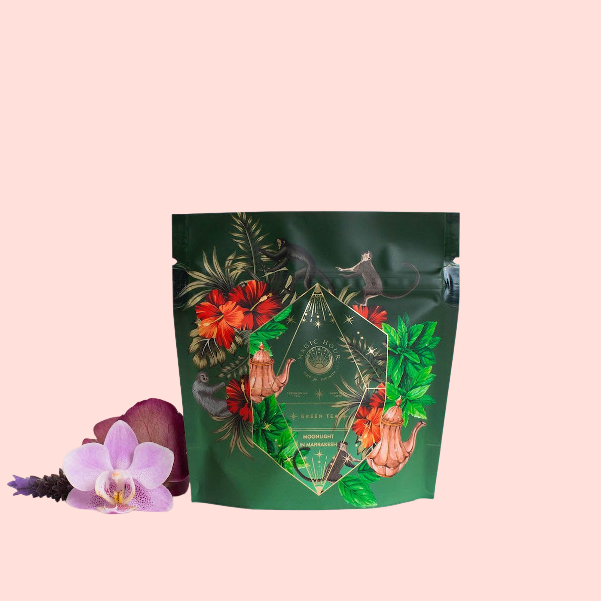 A green, resealable pouch of Moonlight in Marrakesh: Spiced Mint Green Tea, decorated with colorful floral and foliage designs plus gold geometric accents, sits beside a purple orchid and lavender sprigs on a pale pink background.