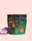 A green, resealable pouch of Moonlight in Marrakesh: Spiced Mint Green Tea, decorated with colorful floral and foliage designs plus gold geometric accents, sits beside a purple orchid and lavender sprigs on a pale pink background.