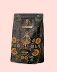 A decorative black tea bag showcases vibrant sunflowers and a woman in orange, with "Magic Hour" and "The Sun" on the front—celebrating The Sun: Energizing Adaptogenic Herbal Tea with Yaupon, Moringa & Turmeric on a light pink background.