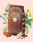 A brown package of Queen of the South Cocoa Detox Tea, featuring gold geometric designs and a sun motif, sits among chicory roots, dandelion leaves and flowers, cinnamon sticks, cocoa beans, and coffee beans on a light pink background.