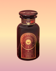 A dark glass bottle with a gold and orange ornate label featuring a sun and geometric designs, labeled "Queen of the South Cocoa Detox Tea," showcases a caffeine-free adaptogenic tea on a warm gradient background.