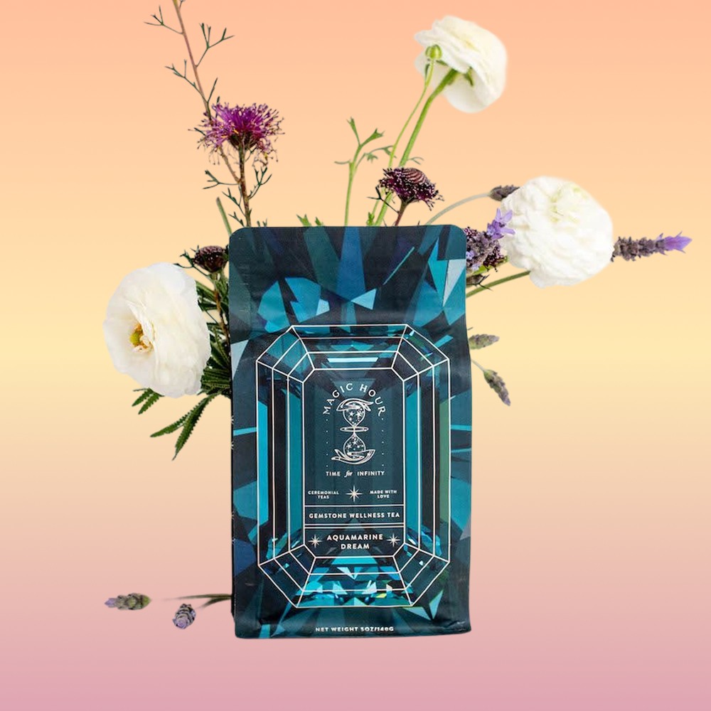 A teal and blue patterned Aquamarine Dream - Soothing Herbal Ayurvedic Adrenal Tonic tea package is displayed before pink and white flowers on a peach gradient background. The label reads "Magic Hour" with a geometric design.
