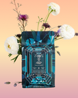 A teal and blue patterned Aquamarine Dream - Soothing Herbal Ayurvedic Adrenal Tonic tea package is displayed before pink and white flowers on a peach gradient background. The label reads "Magic Hour" with a geometric design.
