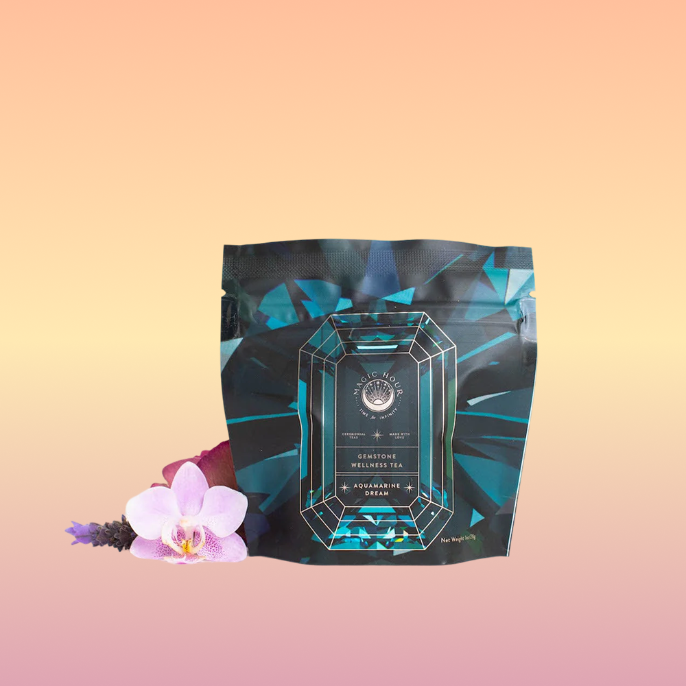 A teal and black "Aquamarine Dream - Soothing Herbal Ayurvedic Adrenal Tonic" tea package sits before a pastel gradient, beside a pink orchid and purple flowers—an adaptogen herbal blend designed for calming sleep support.