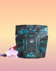 A teal and black "Aquamarine Dream - Soothing Herbal Ayurvedic Adrenal Tonic" tea package sits before a pastel gradient, beside a pink orchid and purple flowers—an adaptogen herbal blend designed for calming sleep support.