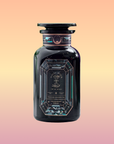 A dark, ornate glass bottle with a decorative label stands upright against a gradient peach-pink background. The label reads “Aquamarine Dream – Soothing Herbal Ayurvedic Adrenal Tonic,” featuring geometric patterns and stylish fonts.