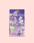 A purple and pink tea package with colorful floral art and a yellow butterfly sits on a light pink background. The label reads "Perfumes of Provence: Lavender Bouquet Tea for Calm Moods and Beautiful Skin.