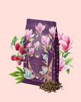 A purple "Breakfast in Paris-Oolong Black Tea Scented with Fruits & Flowers of Secret Parisian Gardens" package, adorned with floral and Paris motifs, is surrounded by magnolias, raspberries, green leaves, and loose tea on a pale pink background.