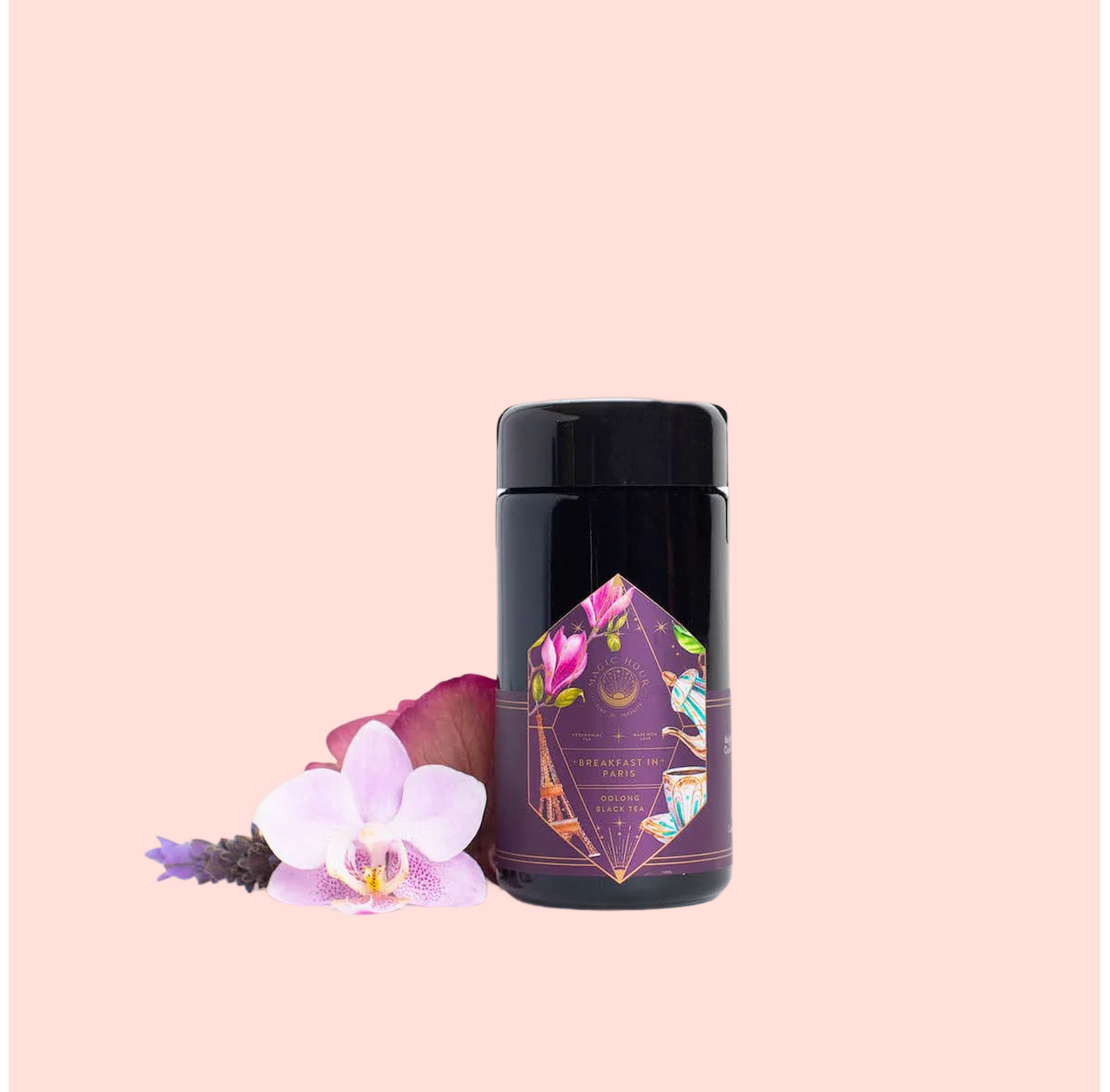 A black cylindrical container of “Breakfast in Paris-Oolong Black Tea Scented with Fruits &amp; Flowers of Secret Parisian Gardens” stands on a pale pink background, adorned with petals and an orchid—suggesting the romance of Parisian gardens.