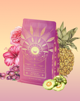 A vibrant pink tea package labeled "Queen of the Tropics: Rosé all Day Alternative Tea" sits among illustrated pineapple, grapes, hibiscus, rose, and Garcinia Cambogia—a refreshing tropical fruit blend and rosé wine alternative.