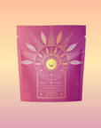 A pink and purple package labeled “Queen of the Tropics: Rosé all Day Alternative Tea” features gold sun, moon, and leaf designs, offering a refreshing tropical fruit tea as a Rosé wine alternative on a peach-yellow background.