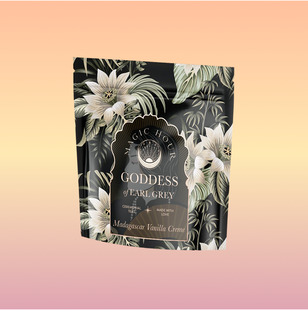 A black floral-patterned packet labeled "Goddess of Earl: Madagascar Vanilla Creme Tea for Soothing Delight & Delicious Decadence" sits centered on a soft peach-to-pink gradient background.