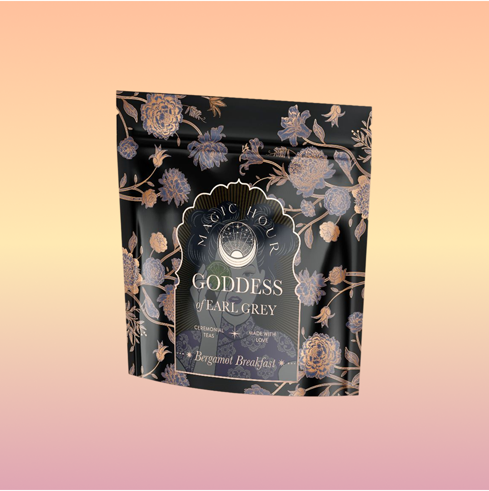 A black and gold floral pouch labeled "Goddess of Earl: Lady Luck Oriental Beauty Oolong Tea" stands against a pink-orange gradient background. The elegant packaging showcases botanical designs and hints of bergamot grapefruit tea.