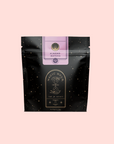 A black, resealable pouch labeled "Almond Matcha Green Tea for Joy" with gold stars, lavender accents, and a circular hourglass logo is shown on a light pink background, highlighting this immune-boosting organic tea.