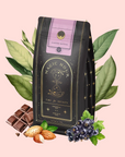 A black bag of Almond Matcha Green Tea for Joy with gold stars and an hourglass design sits on a light pink background, surrounded by green leaves, chocolate pieces, almonds, blackcurrants, and mint.