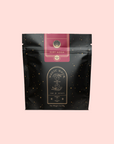 A black and gold pouch labeled "Magic Hour" with a celestial design and "Ruby Moon™ : Hibiscus Elderberry Tea" on a light pink background. The caffeine-free tea packaging has small gold dots and a decorative emblem at the top.