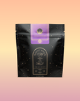 A black resealable tea pouch with gold star patterns, labeled "Lucid Dreams: Tulsi-Turmeric Herbal Tea for Sleep & Calm" in purple and gold, is shown against a pastel peach-pink gradient—an inviting choice for restful, caffeine-free relaxation.