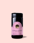 A black jar labeled "Almond Matcha Green Tea for Joy" with pink and gold accents stands against a light pink background. The label shows the net weight as 1 oz (28g) and highlights its immune-boosting benefits.