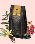 A black and gold bag of "Bohemian Breakfast Vanilla Puerh Black Tea for Digestion & Energy" with celestial designs is surrounded by tea leaves, vanilla flower and pod, pomegranate, and dark berries on a light pink background.